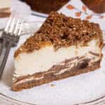 A slice of the Apple Crisp Cheesecake sits on a tan oplate with a fork on the plate and a napkin in the background.