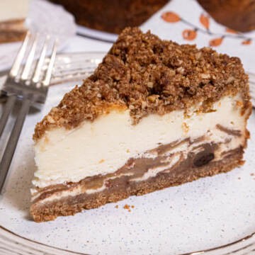 A slice of the Apple Crisp Cheesecake sits on a tan oplate with a fork on the plate and a napkin in the background.