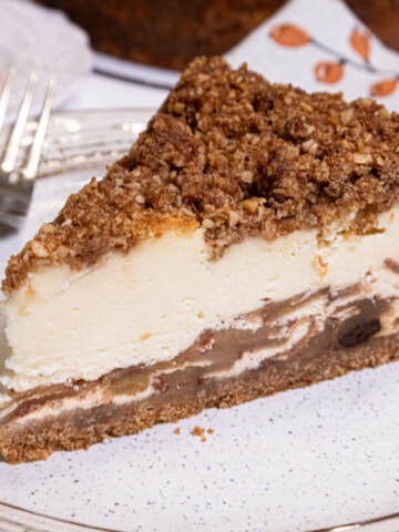 A slice of the Apple Crisp Cheesecake sits on a tan oplate with a fork on the plate and a napkin in the background.