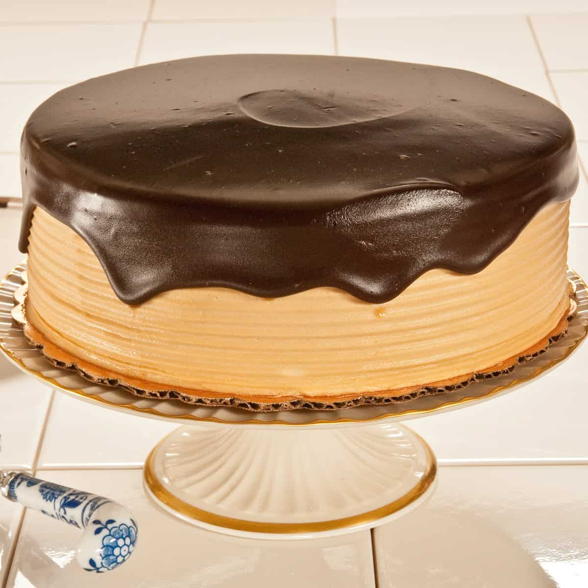An Espresso  Fudge Cake with its beige frosting and a dark chocolate ganache dripping down the sides sits on an ivory cake plate with a server next to it.
