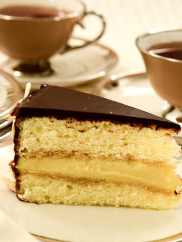 A slice of Boston Cream Pie on a plate with cups of tea in the background,