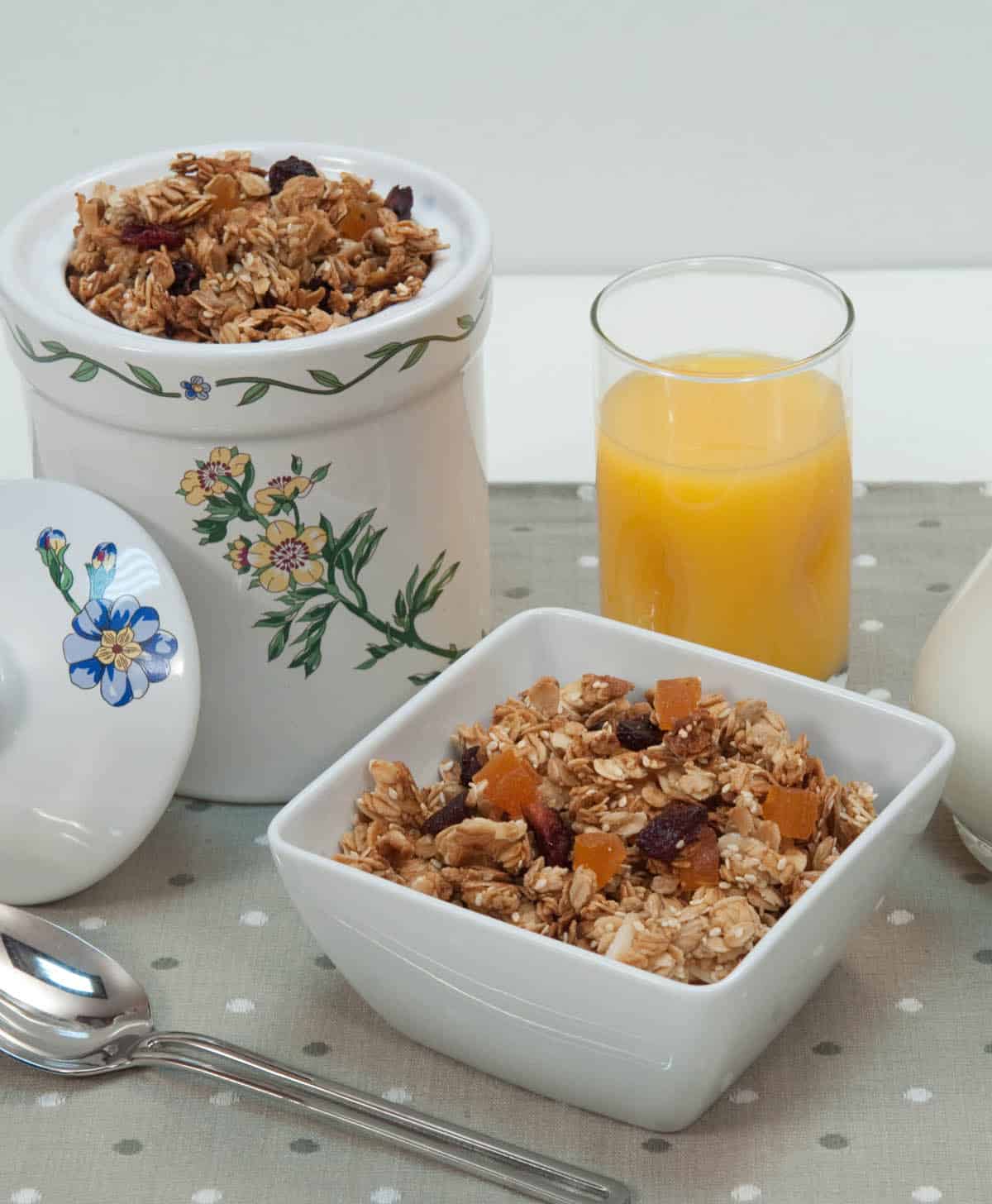 A canister of Granola along with a bowl of granola and orange juice are featured in this photo.