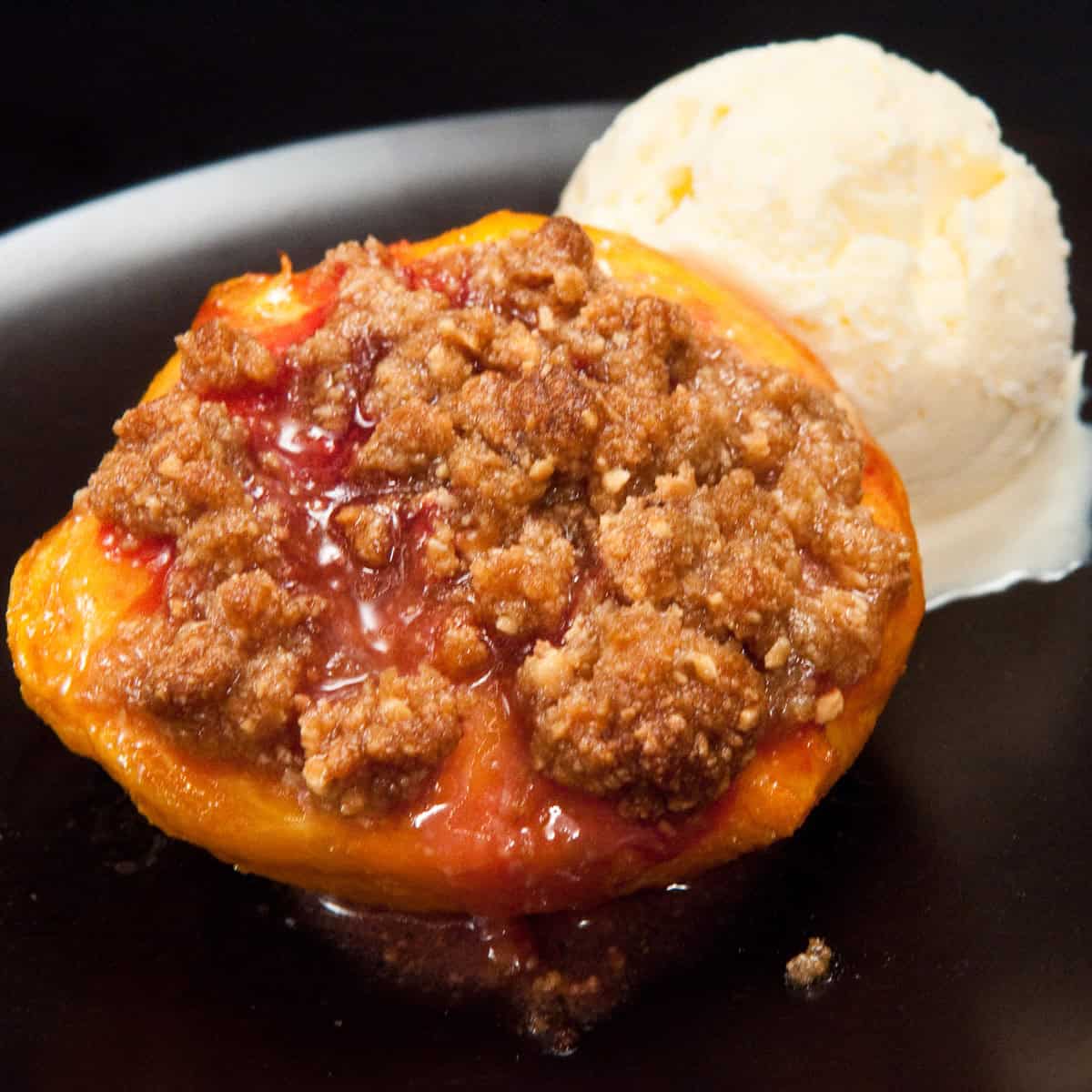 A Roasted Peach with Amaretti Crumble sits on a black plate with a scoop of vanilla ice cream .