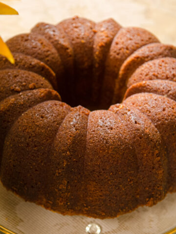 A warm brown Lemon Rum Bundt Cake on a clear glass plate with petals of a yellow flower.