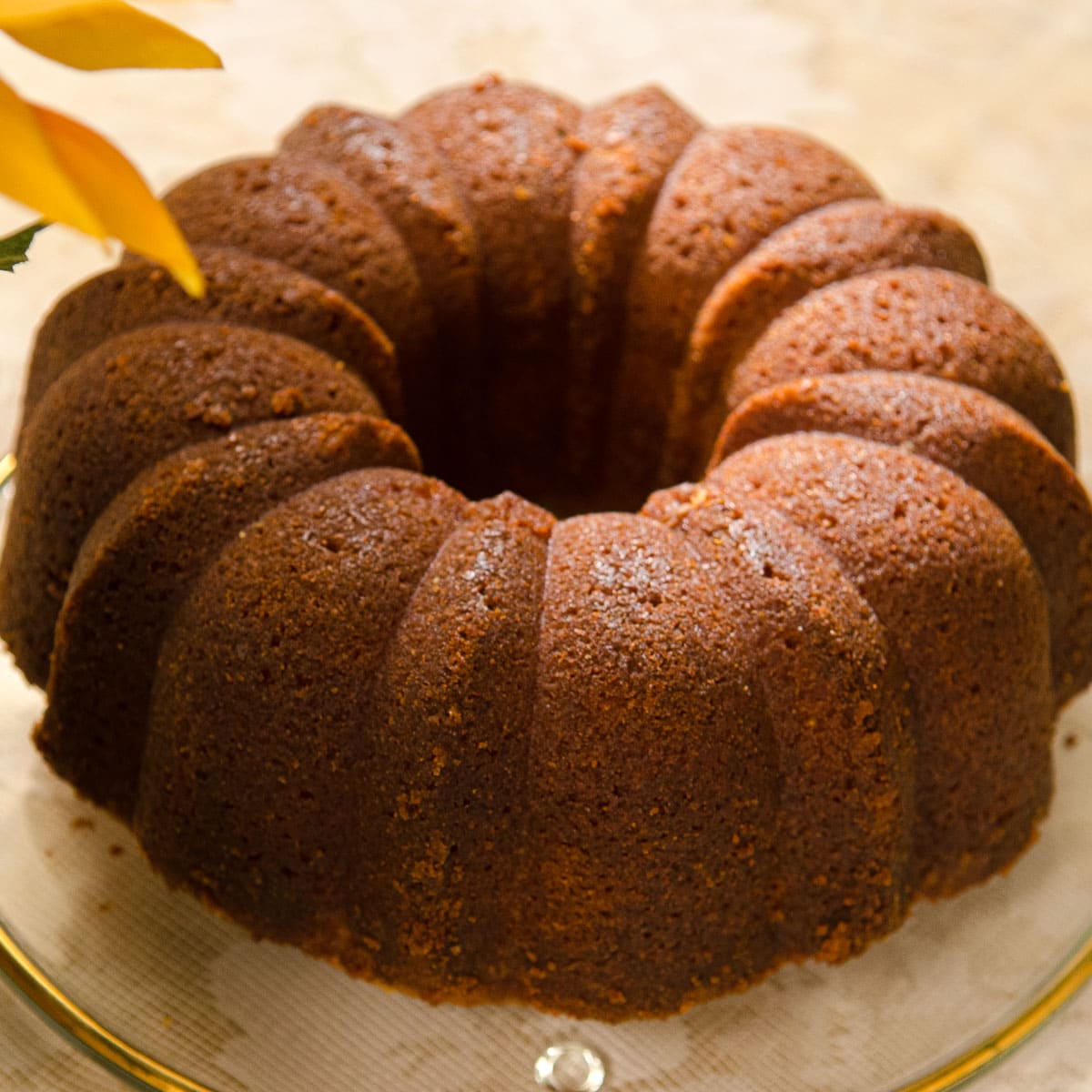 A warm brown Lemon Rum Bundt Cake on a clear glass plate with   petals of a yellow flower.