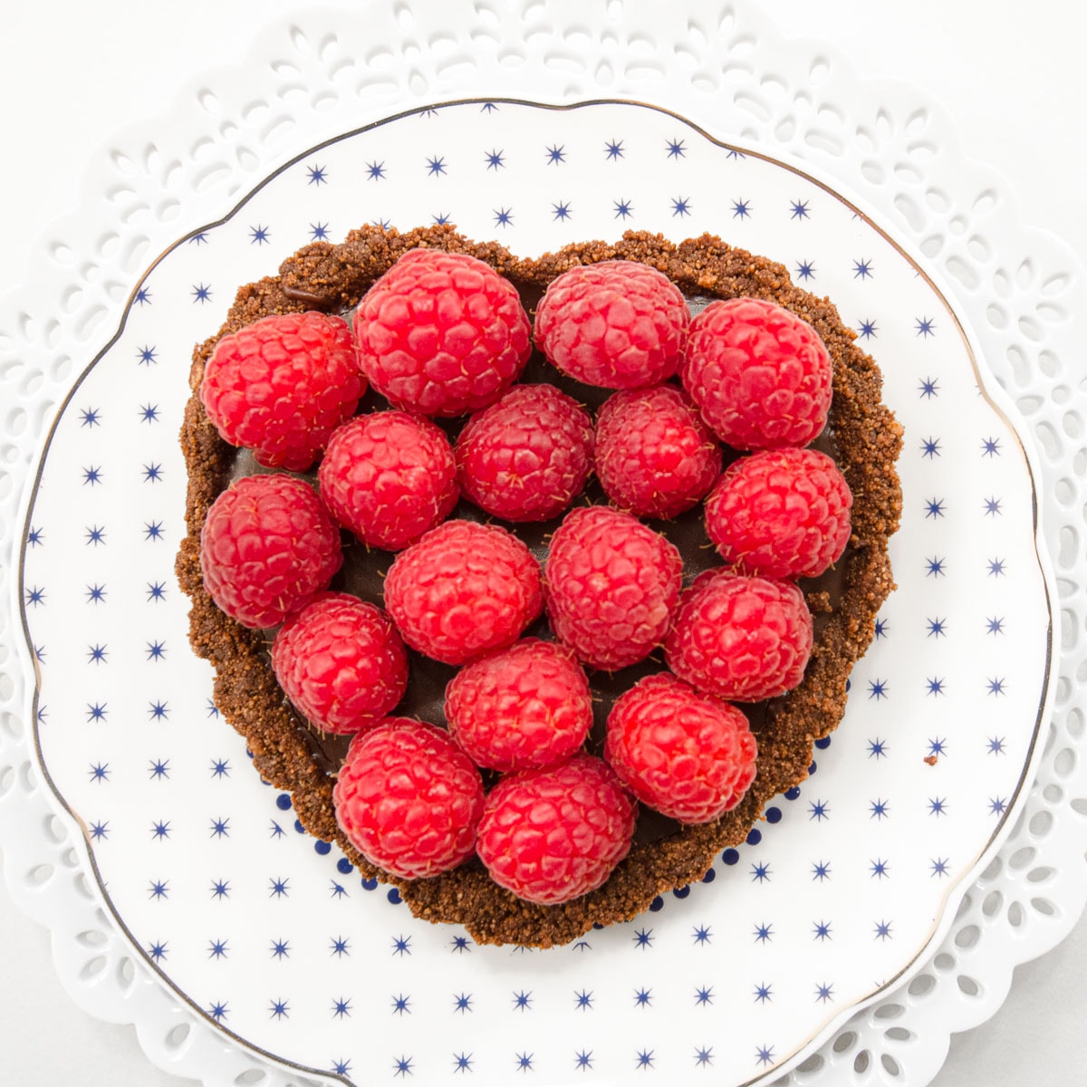 A Chocolate Raspberry Truffle Tart sits on a gold edged white plate with blue dots on top of a lace edged white plate.