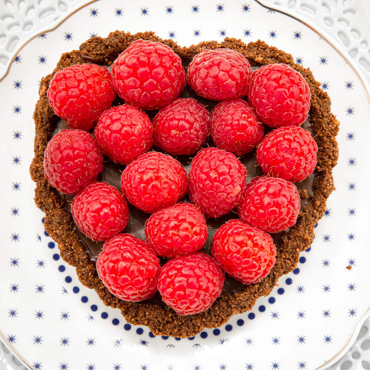 A heart shaped tart filled with truffle filling and topped with fresh raspberries on a white plate with blue dots.