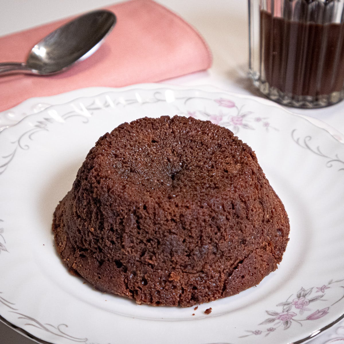 A Warm Lava Cake sits on an antique plate with a spoon and coral napking and Bailey Sauce in the background.