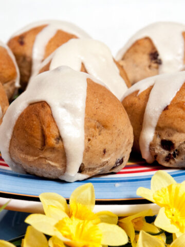 A photo of Hot Cross Buns on a colored cake plate surrounded by yellow daffodils.