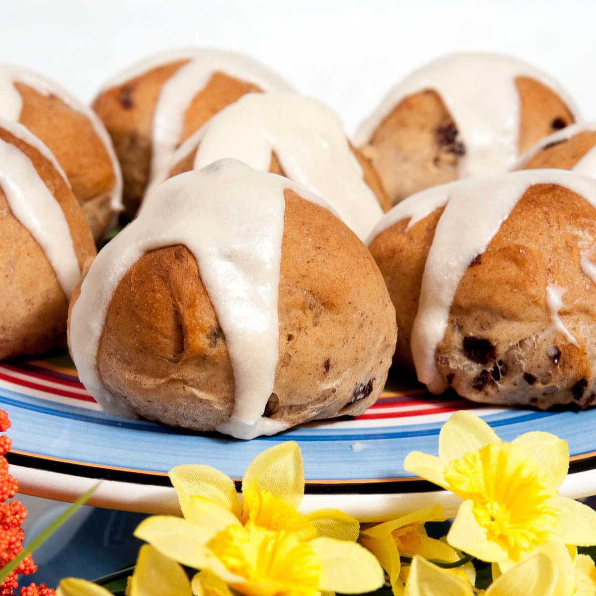 A photo of Hot Cross Buns on a colored cake plate surrounded by yellow daffodils.