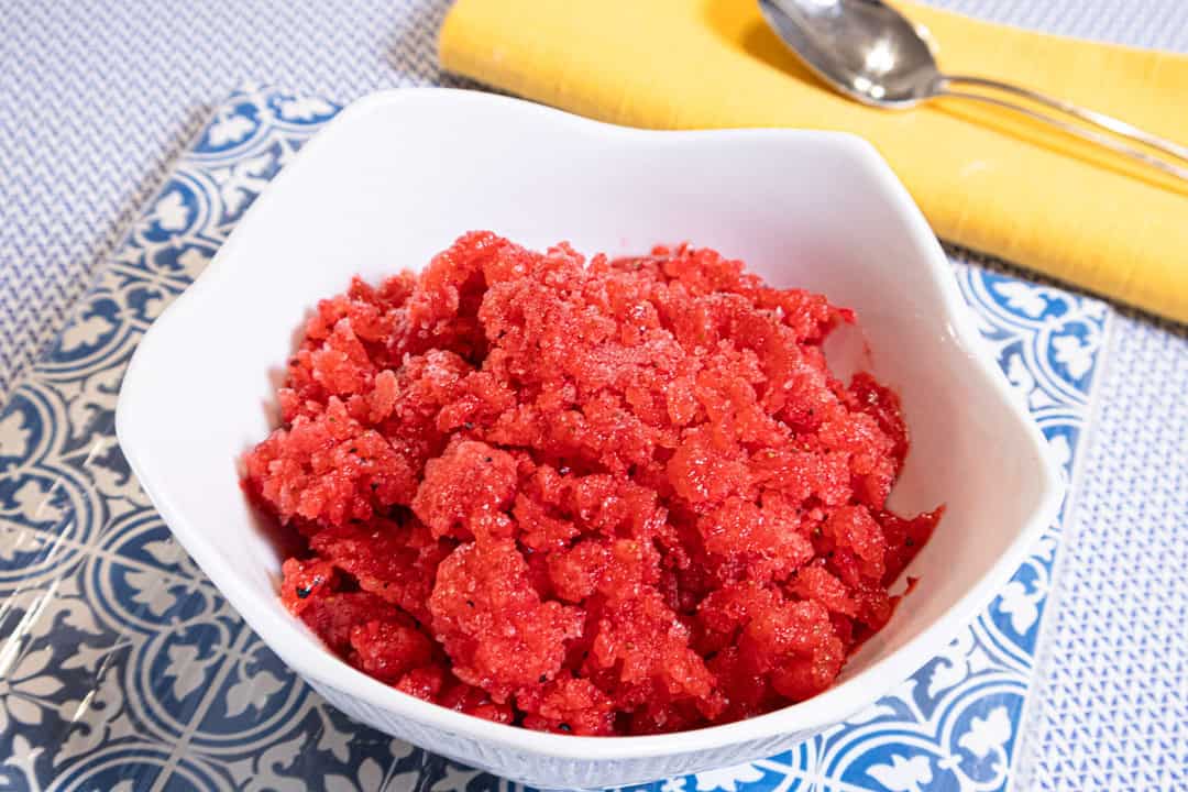 Strawberry Granita sists in a white bowl on a blue and white background with a yellow napkin and spoon .