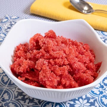 Strawberry Granita sists in a white bowl on a blue and white background with a yellow napkin and spoon .