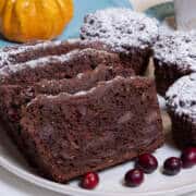 Several slices of Chocolate Cranberry Bread sists on a cream colored plate surrounded by Muffins, cranberries, a teal colored napkin, cup and saucer and a miniature pumpkin gourd.