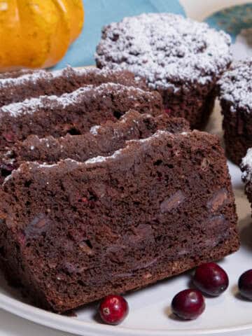 Several slices of Chocolate Cranberry Bread sists on a cream colored plate surrounded by Muffins, cranberries, a teal colored napkin, cup and saucer and a miniature pumpkin gourd.