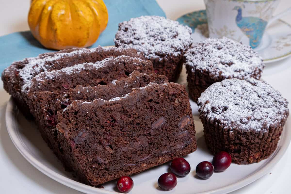 Several slices of Chocolate Cranberry Bread sists on a cream colored plate surrounded by Muffins, cranberries, a teal colored napkin, cup and saucer and a miniature pumpkin gourd.