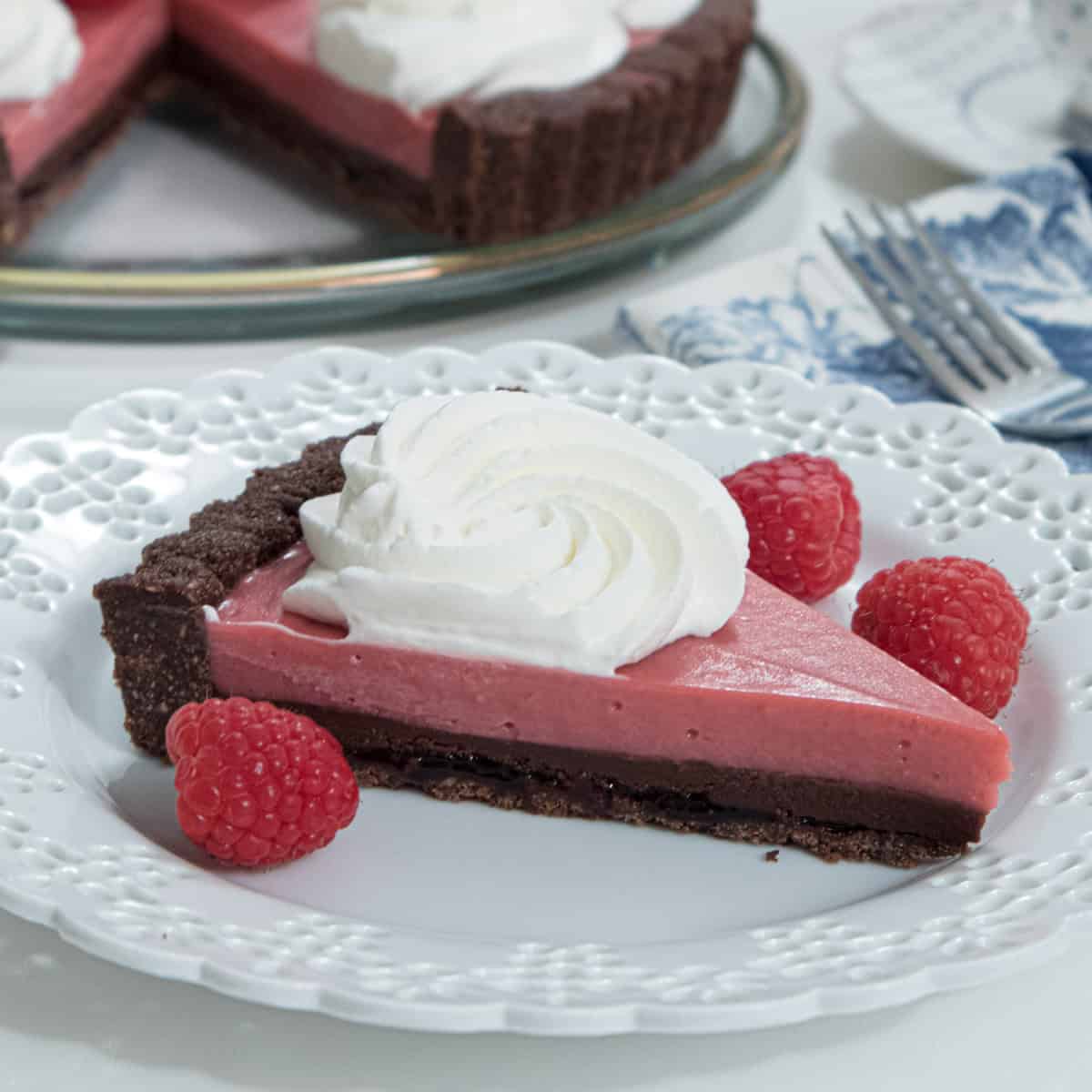 A slice of the Chocolate Truffle Raspberry Curd Tart sits on a lace edged white plate with a few red raspberries and the tart in the background.