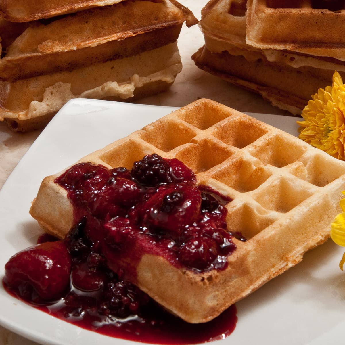 A Belgium waffle on a plate with a fruit compote and stacks of waffles in the background.