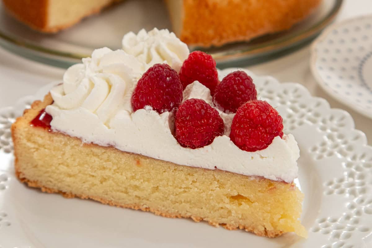 A lice of cake with the whipped cream and raspberries on a lace edged plate with the cake in the background.