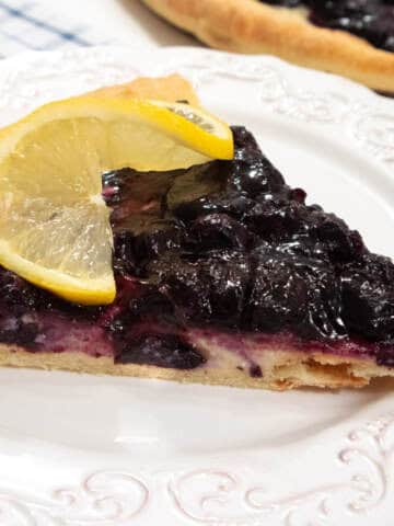 A slice of the blueberry Pizza sits on a white plate with a lemon slice and the pizza in the background.