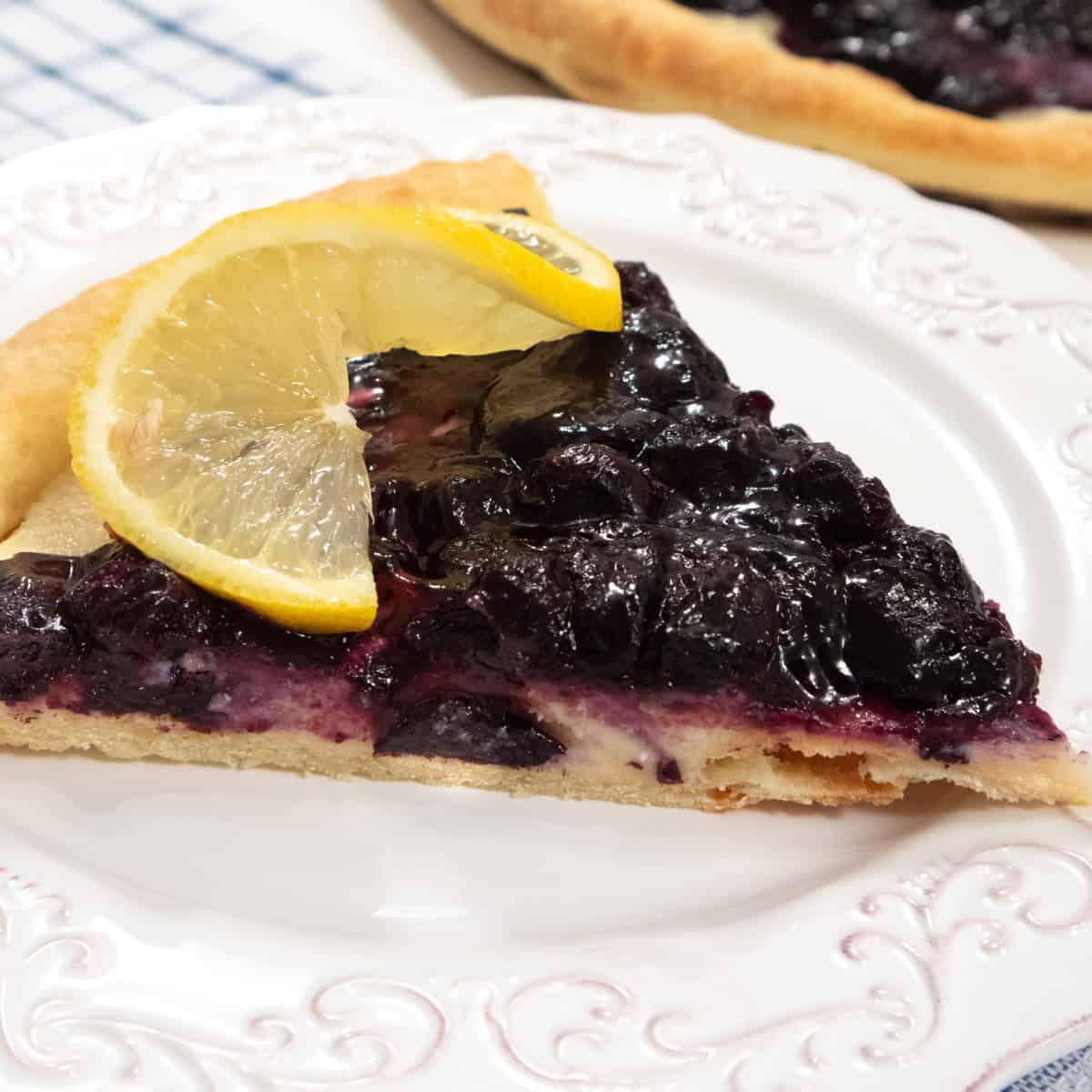 A slice of the blueberry Pizza sits on a white plate with a lemon slice and the pizza in the background.