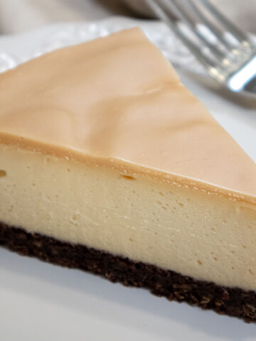 A white chocolate glaze on top of the Bailey's Cheesecake sitting on a dark chocolate crust. The cheesecake sits on a white plate with a fork on the plate.