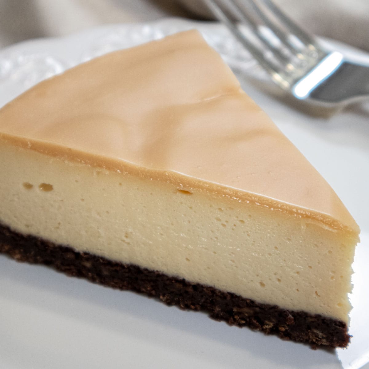 A white chocolate glaze on top of the Bailey's Cheesecake sitting on a dark chocolate crust.  The cheesecake sits on a white plate with a fork on the plate.