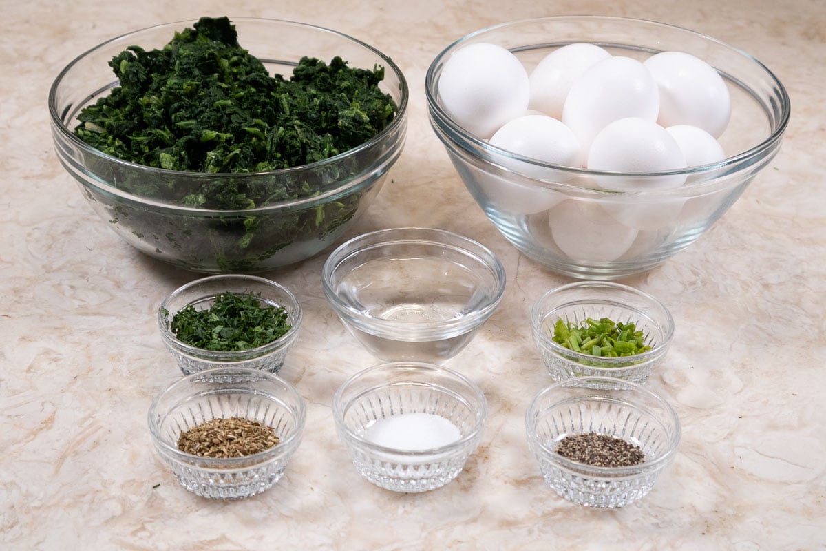 The ingredients for the omelet include eggs, parsley, water, chives, salt, pepper, and oregano.  The spinach in the photo is not used in the omelet.