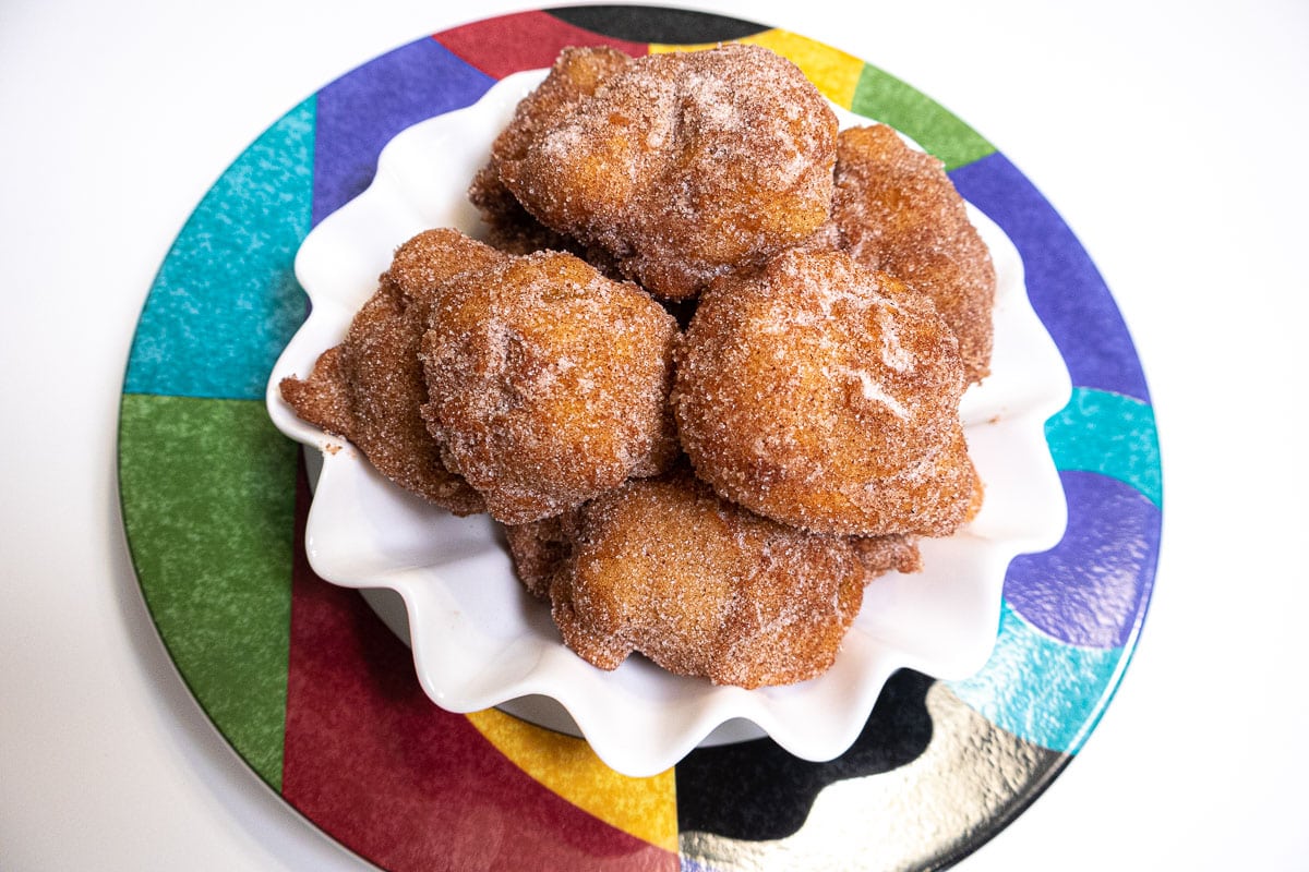 Apple Fritters are in a white bowl with rippled edges sitting on a multi-colored plate.