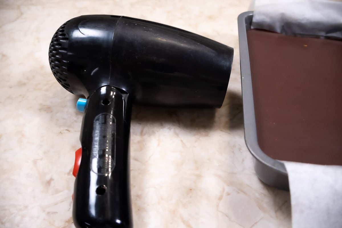 A hair dryer is held close to the pan with the No Bake Cookie Bar to heat the side so the cookie can release cleanly.