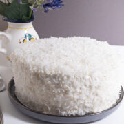 The finished Coconut Cream Cake sits on a gray plate with a flower vase in the background,