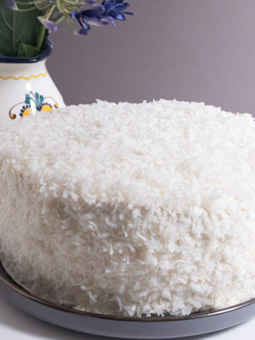 The finished Coconut Cream Cake sits on a gray plate with a flower vase in the background,