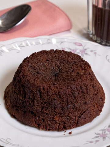 A Warm Lava Cake sits on an antique plate with a spoon and coral napking and Bailey Sauce in the background.