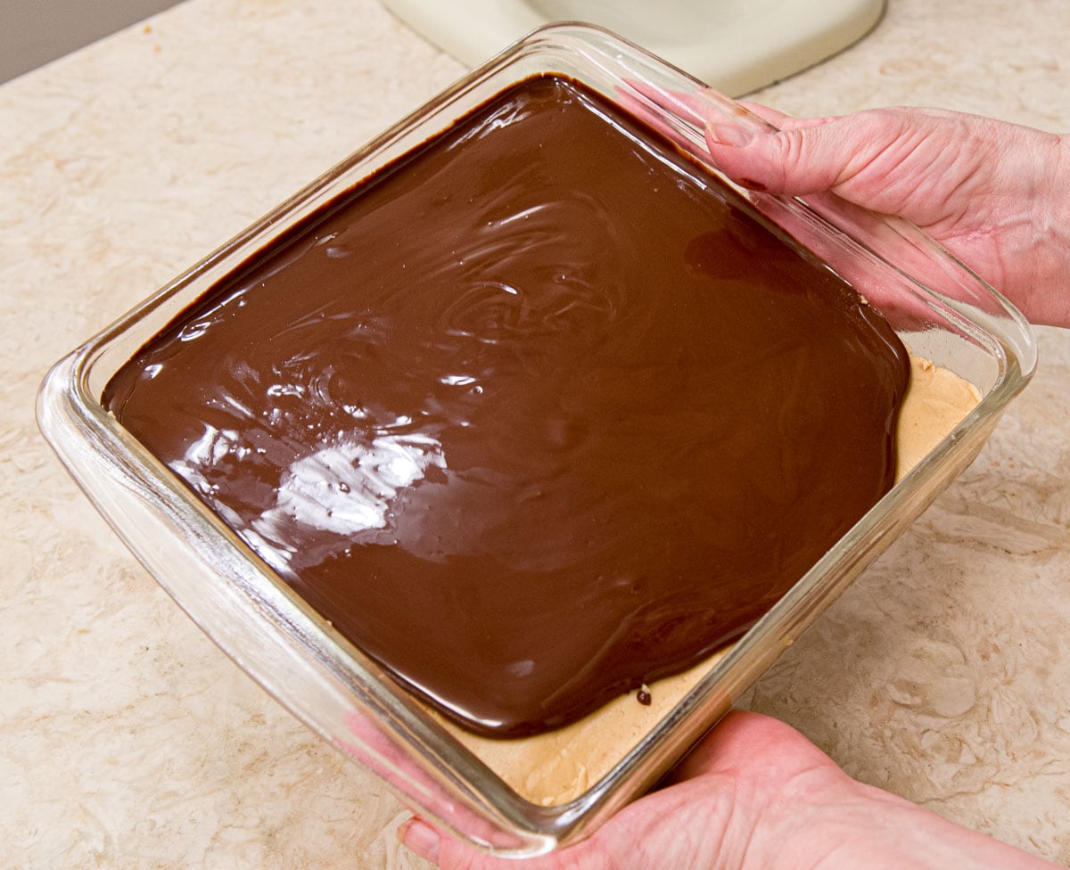 The topping has been poured over the peanut butter layer and is being tilted to spread it.