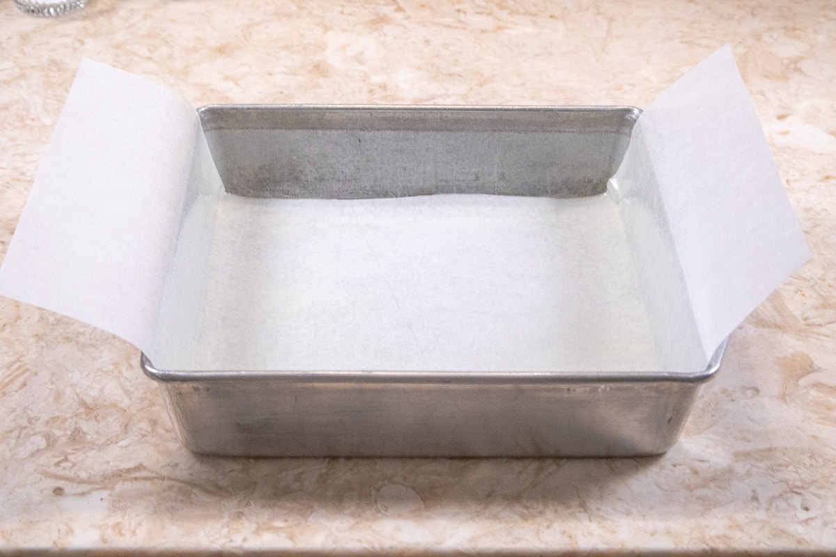 A 9x9" pan is lined with parchment paper allowing overhang on two sides.