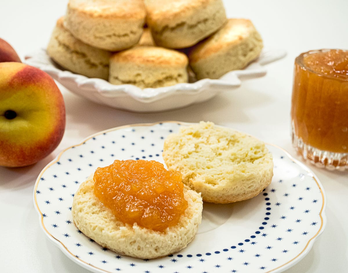 A scone on a plate is sliced open and the jam is on one side with a plate of scones behind it and a container of jam beside the plate.