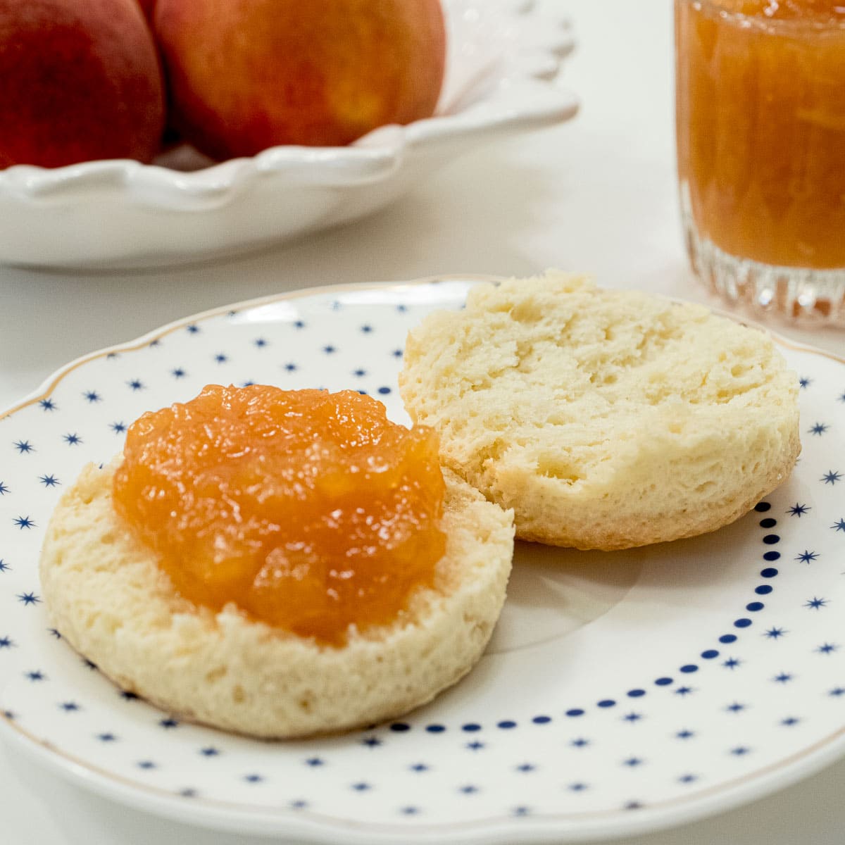 Peach Jam sits on a biscuit with a bowl of peaches and a container of jam behind it.
