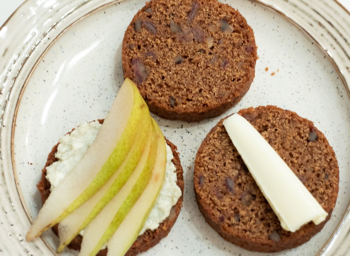 Three rounds of date nut bread are on a plate.  One is plain, one has bleu cheese and pear slices and the third has butter on it. 