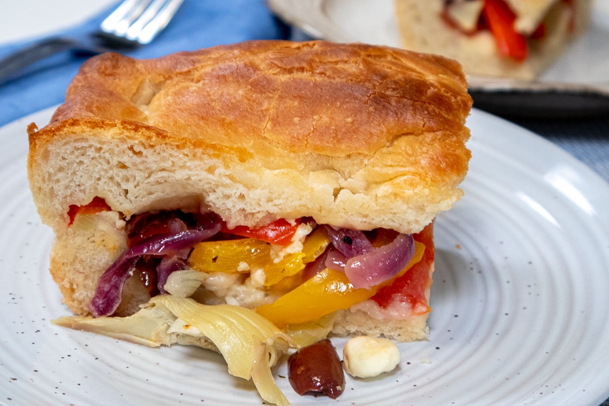 A hefty slice of focaccia sits on a light gray plate with veggies tumbling out.