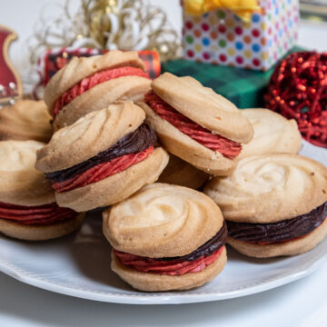 A plate of Viennese Whirls with strawberry and chocolate filling in front of gift boxes.