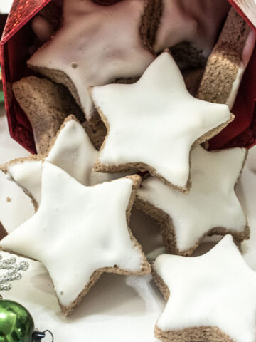 White, star shaped cinnamon Zimtsterne cookies are flowing fro a container surounded by holiday ornaments.