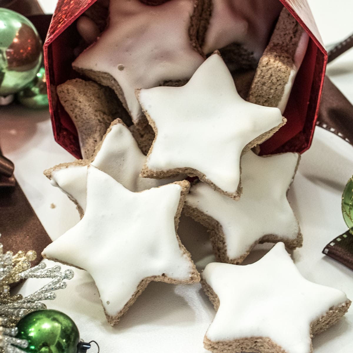 White, star shaped cinnamon Zimtsterne cookies are flowing fro a container surounded by holiday ornaments.  