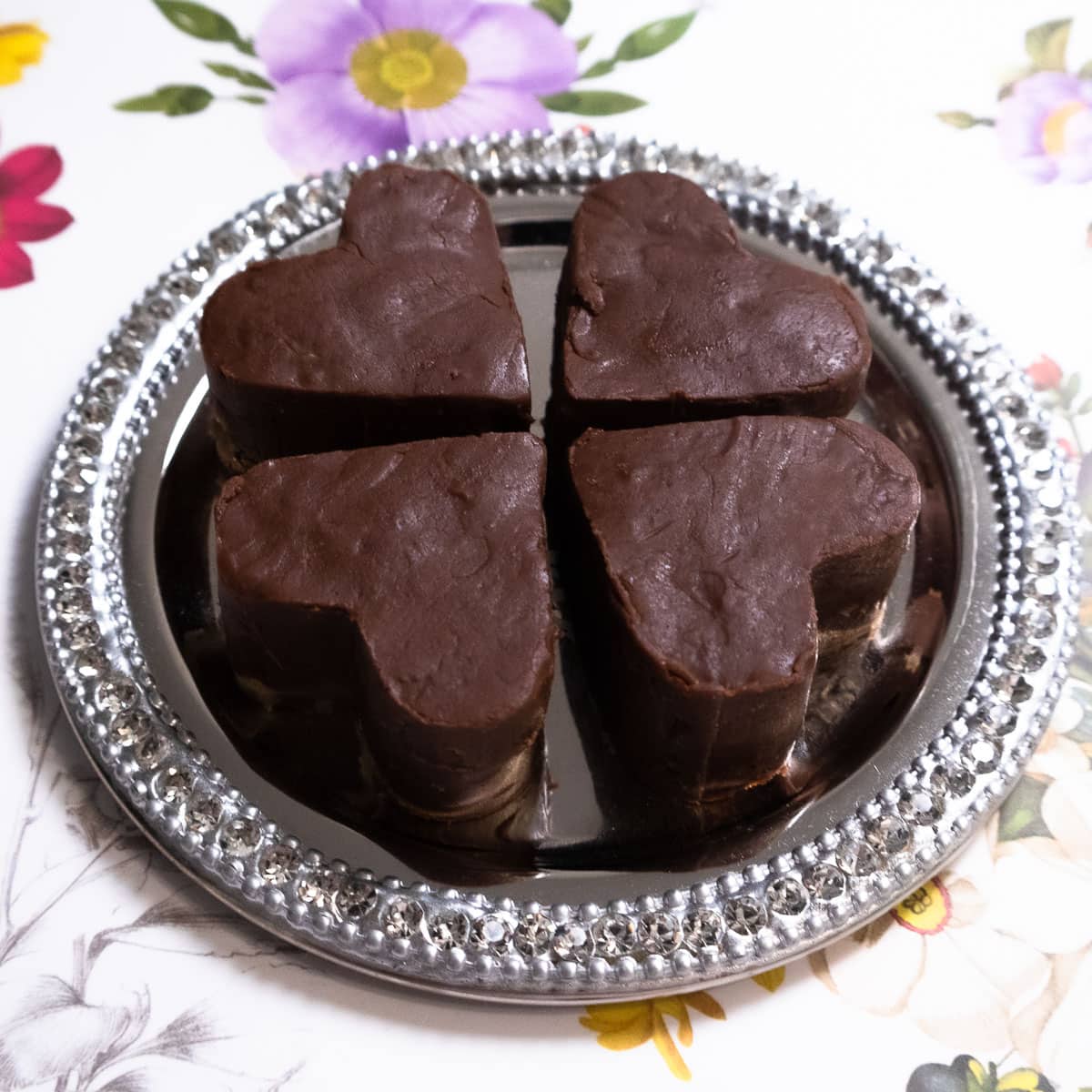 Four chocolate hearts sit on a silver plate in front of a floral setting.