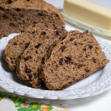 Slices of Traditional Irish Soda bread are on a lace edged white plate with the loaf and butter behind.