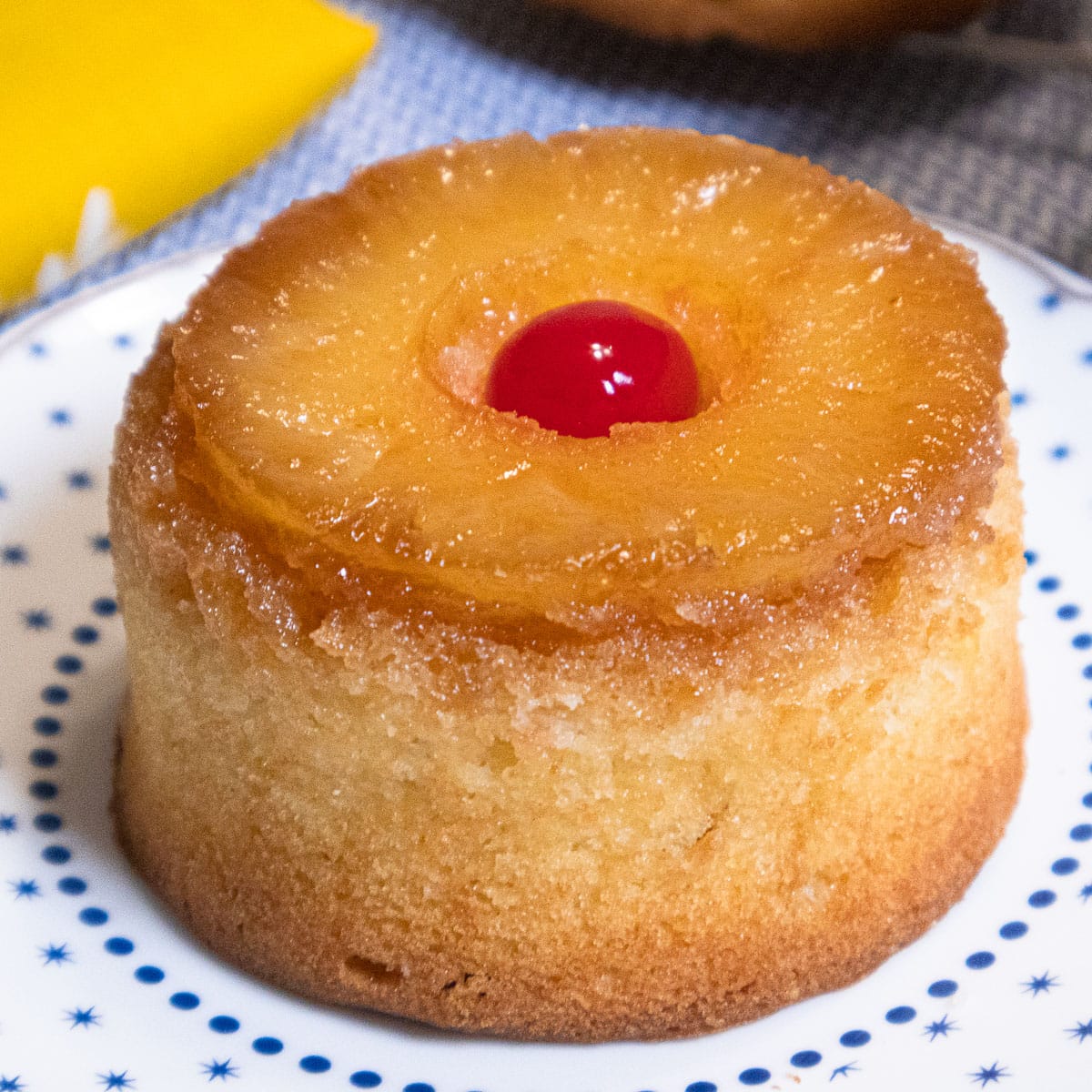 This Mini Pineapple Upside Down Cake features a pineapple slice with a cherry in the middle on top of a yellow cake sitting on a blue and white plate.