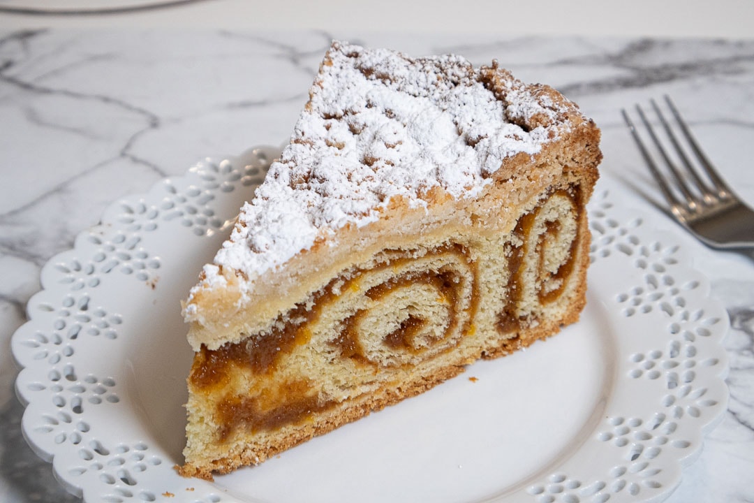 This picture shows a slice of the coffee cake on a white, lace edged plate on a marble counter.
