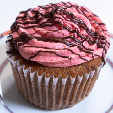 A lone Banana Split Cupcake with its vibrant pink strawberry buttercream and drizzled with chocolate sits on a pink rimmed plate.
