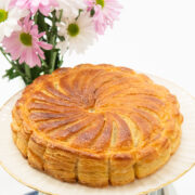 A golden brown,many layered French pastry, Pithiviers filled with pastry cream sits on a cream cake plate with pink and white daisies in the background.