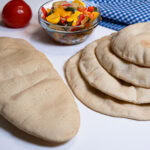 This photo shows a variety of round pizza crusts and a flatbread crust with a tomato and sauteed veggies in the background.