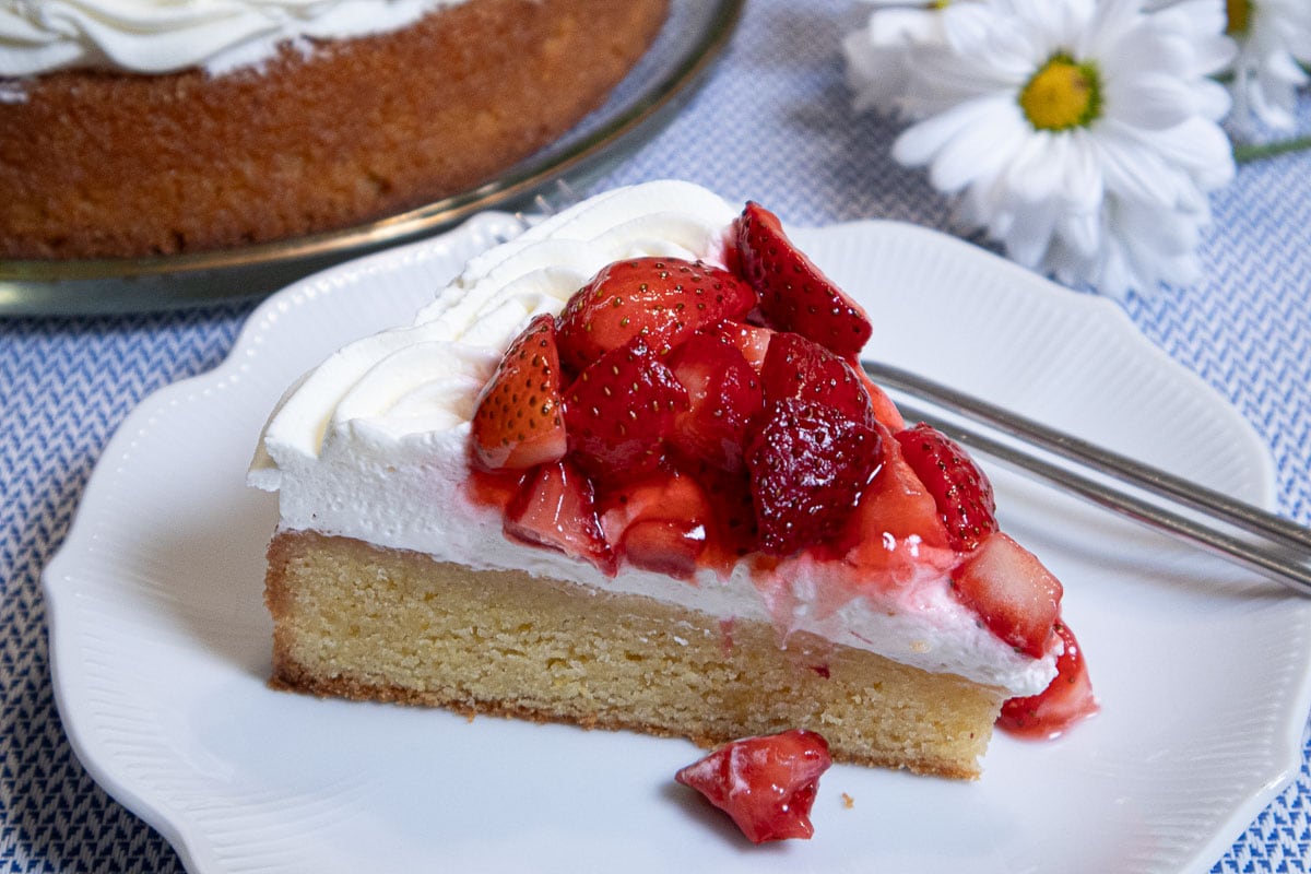This photo is of a slice of the Lemon Strawberry Cake on a white plate with a daisy in the background and the cake on a serving plate.