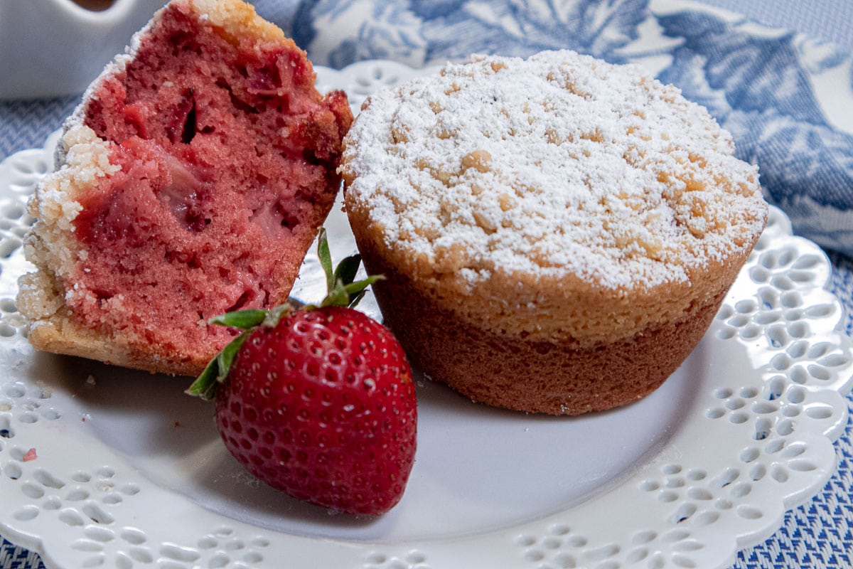 A whole muffin sits next to an open muffin with a strawberry on a white plate with a blue and white napkin in the background.